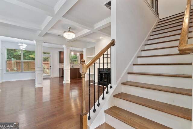 stairway featuring ornate columns, coffered ceiling, beam ceiling, hardwood / wood-style flooring, and a chandelier