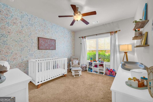 bedroom featuring light carpet, a ceiling fan, visible vents, a nursery area, and wallpapered walls