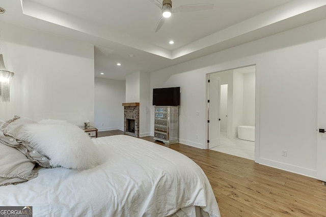 bedroom with a brick fireplace, light hardwood / wood-style flooring, a raised ceiling, and ensuite bathroom