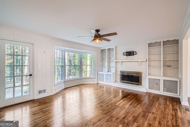 unfurnished living room featuring a brick fireplace, ceiling fan, crown molding, wood-type flooring, and built in features