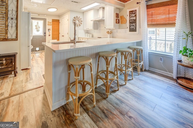 kitchen with kitchen peninsula, wall chimney range hood, light hardwood / wood-style floors, white cabinetry, and a breakfast bar area