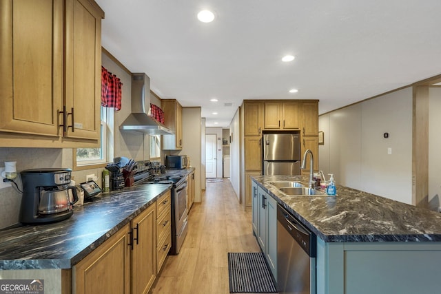 kitchen with wall chimney range hood, sink, an island with sink, appliances with stainless steel finishes, and light hardwood / wood-style floors