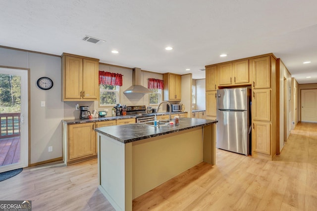 kitchen with sink, stainless steel appliances, wall chimney range hood, light hardwood / wood-style flooring, and a kitchen island with sink