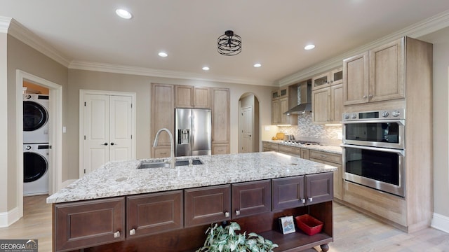 kitchen with sink, wall chimney range hood, stacked washer / drying machine, a kitchen island with sink, and appliances with stainless steel finishes