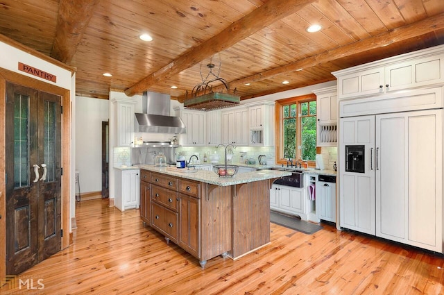 kitchen featuring light stone counters, wall chimney exhaust hood, paneled built in refrigerator, a center island with sink, and white cabinets