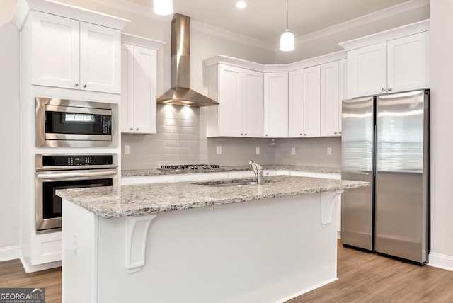 kitchen with white cabinets, appliances with stainless steel finishes, wall chimney exhaust hood, and an island with sink