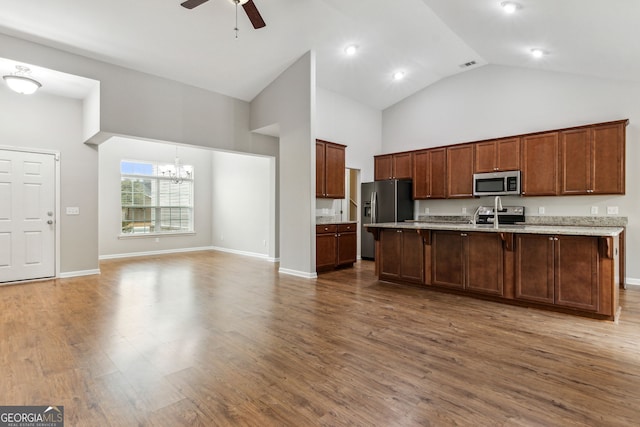 kitchen with light stone countertops, an island with sink, wood-type flooring, ceiling fan with notable chandelier, and appliances with stainless steel finishes