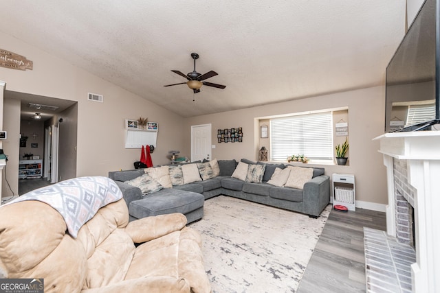 living room featuring ceiling fan, wood finished floors, visible vents, vaulted ceiling, and a brick fireplace