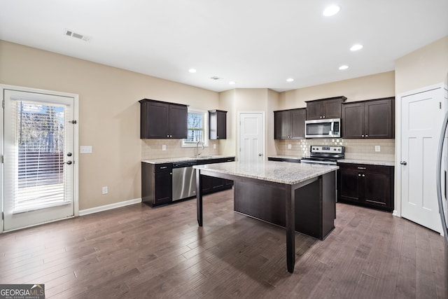 kitchen featuring appliances with stainless steel finishes, a kitchen island, dark brown cabinetry, dark wood-type flooring, and light stone counters