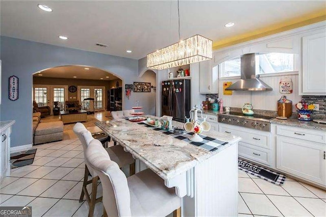 kitchen featuring a kitchen bar, wall chimney exhaust hood, black fridge with ice dispenser, stainless steel gas cooktop, and white cabinets