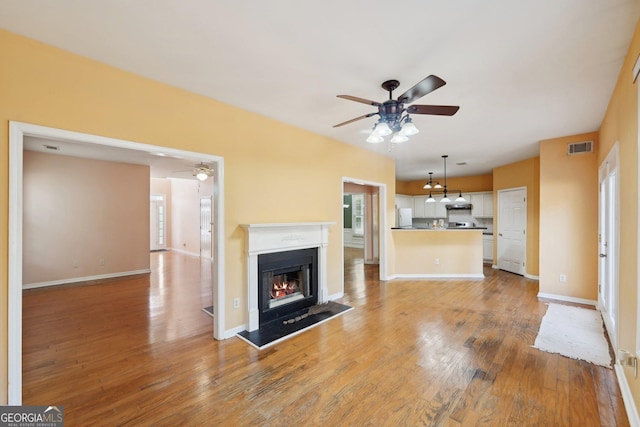 unfurnished living room featuring ceiling fan, a warm lit fireplace, visible vents, baseboards, and light wood-style floors