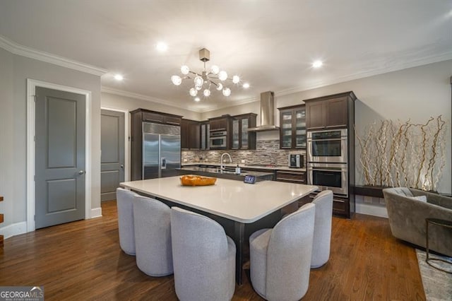 kitchen with appliances with stainless steel finishes, backsplash, a kitchen island with sink, dark brown cabinetry, and wall chimney exhaust hood