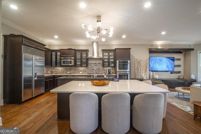 kitchen featuring pendant lighting, wall chimney range hood, stainless steel appliances, dark brown cabinetry, and a center island with sink