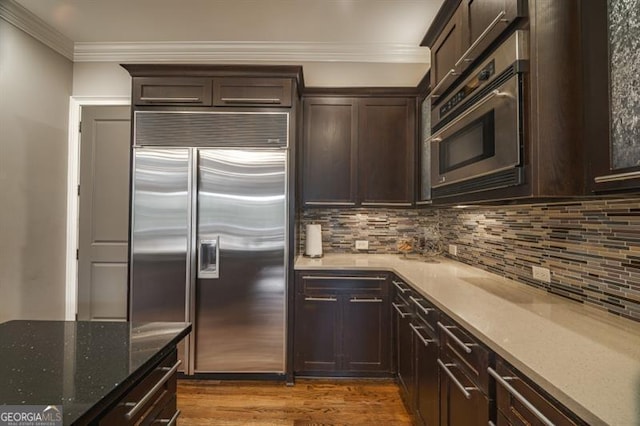 kitchen with crown molding, built in appliances, dark brown cabinetry, light stone countertops, and decorative backsplash