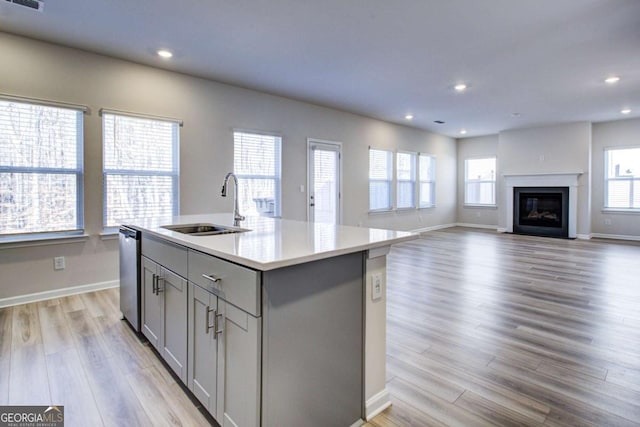 kitchen with gray cabinetry, a center island with sink, sink, stainless steel dishwasher, and light wood-type flooring