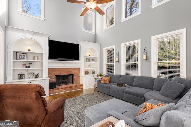 living room with a towering ceiling, a brick fireplace, ceiling fan, built in features, and hardwood / wood-style floors