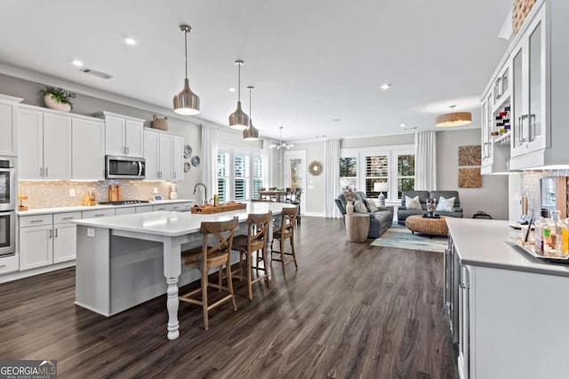 kitchen with a wealth of natural light, stainless steel appliances, decorative light fixtures, white cabinetry, and an island with sink