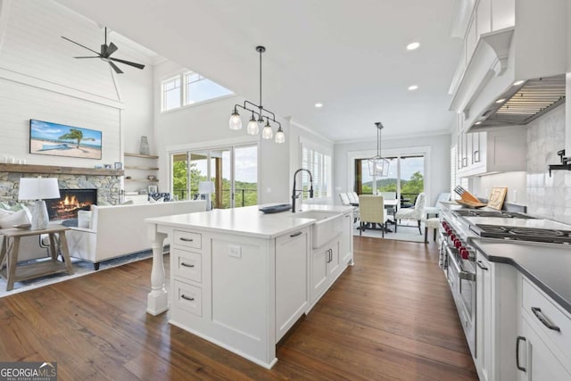 kitchen featuring wall chimney exhaust hood, white cabinets, a stone fireplace, hanging light fixtures, and an island with sink