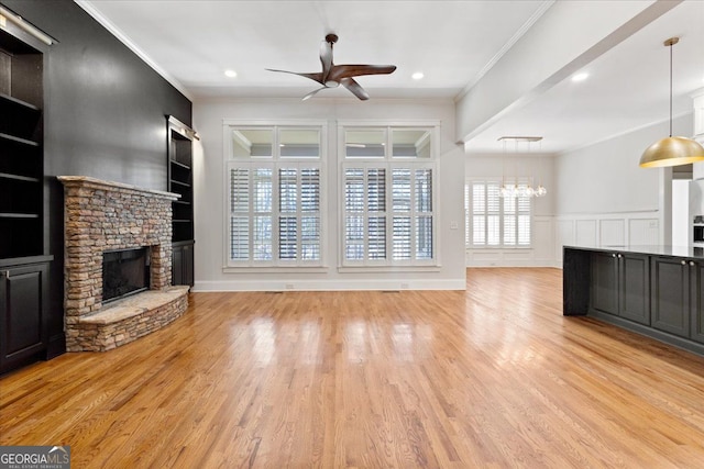 unfurnished living room with built in shelves, a stone fireplace, light hardwood / wood-style flooring, crown molding, and ceiling fan with notable chandelier