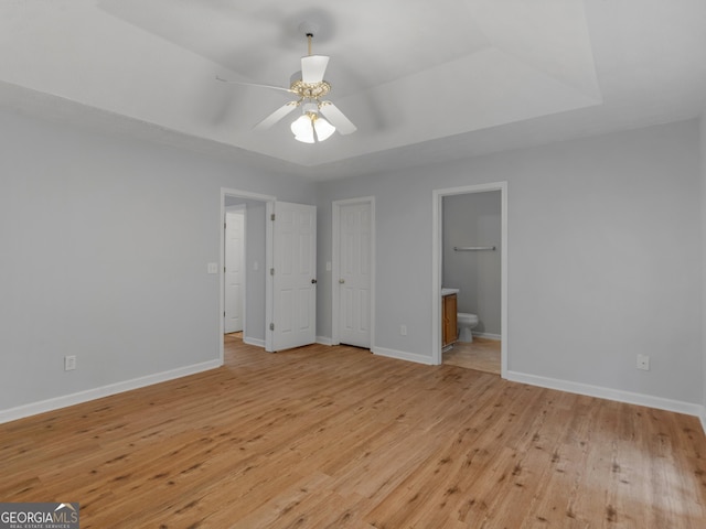 unfurnished bedroom featuring ceiling fan, light hardwood / wood-style floors, a tray ceiling, and ensuite bath