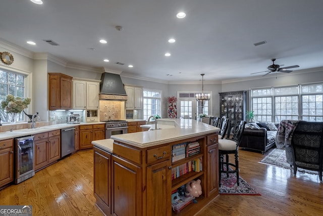 kitchen featuring appliances with stainless steel finishes, custom exhaust hood, a kitchen island with sink, decorative light fixtures, and wine cooler