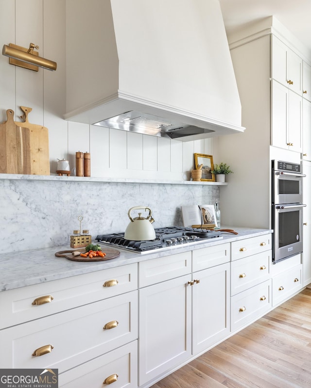 kitchen with custom exhaust hood, tasteful backsplash, appliances with stainless steel finishes, light hardwood / wood-style floors, and white cabinets