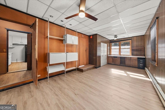 interior space with wood walls, a drop ceiling, a baseboard heating unit, and ceiling fan