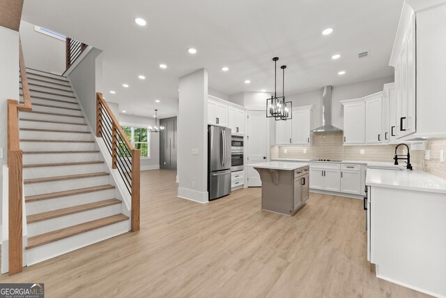 kitchen with appliances with stainless steel finishes, wall chimney exhaust hood, white cabinets, a center island, and hanging light fixtures