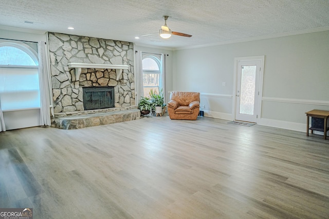 living room with ceiling fan, a stone fireplace, ornamental molding, a textured ceiling, and light wood-type flooring