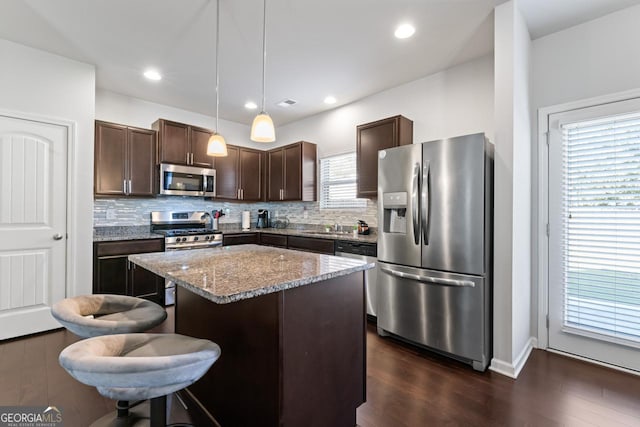 kitchen with dark brown cabinetry, light stone countertops, a center island, hanging light fixtures, and appliances with stainless steel finishes