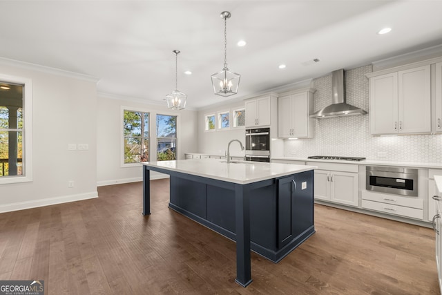 kitchen featuring wall chimney range hood, sink, white cabinetry, hanging light fixtures, and an island with sink