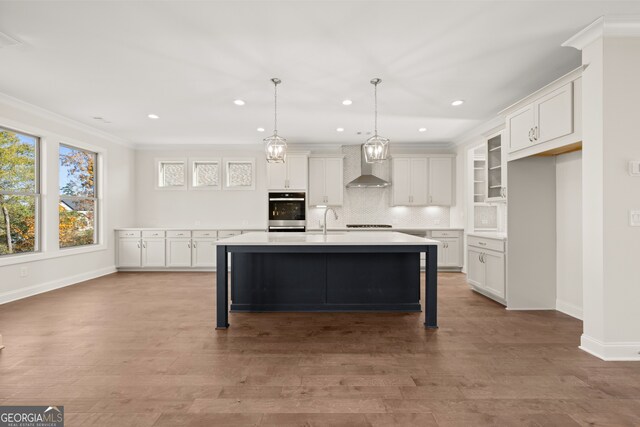 kitchen featuring stainless steel oven, decorative light fixtures, a center island with sink, wall chimney range hood, and white cabinets