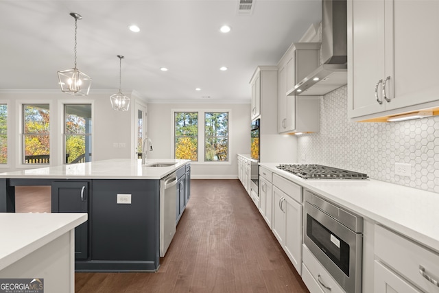 kitchen featuring wall chimney range hood, sink, appliances with stainless steel finishes, an island with sink, and decorative light fixtures