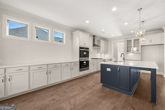 kitchen with pendant lighting, wall chimney range hood, a kitchen island with sink, backsplash, and white cabinets