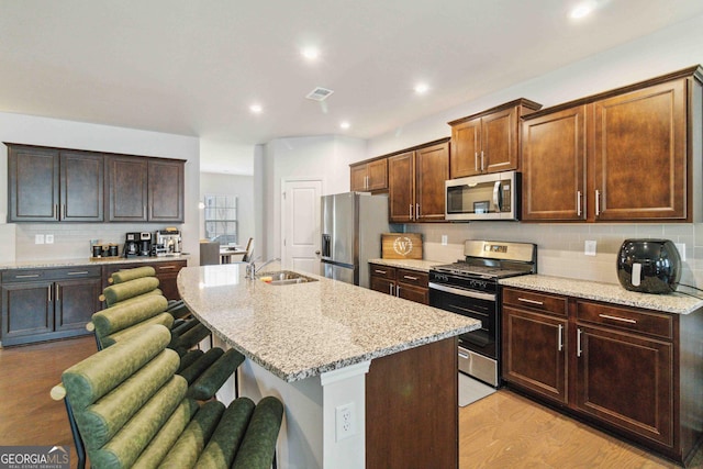 kitchen with light stone counters, light hardwood / wood-style flooring, a kitchen island with sink, a breakfast bar, and appliances with stainless steel finishes