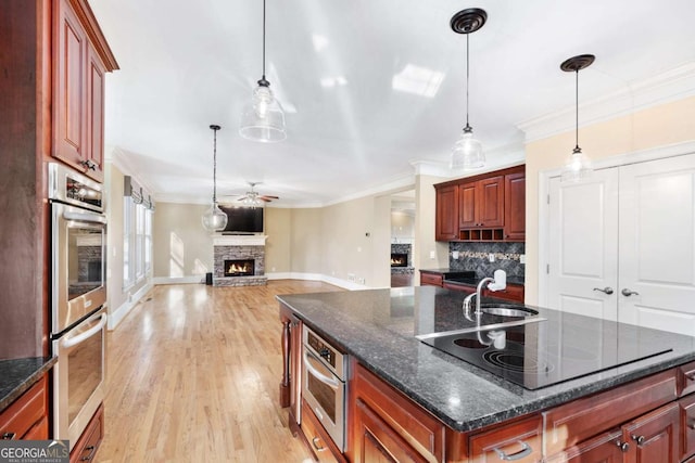 kitchen with a center island with sink, a stone fireplace, ceiling fan, black electric cooktop, and stainless steel double oven