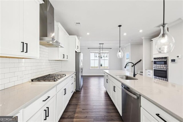 kitchen with appliances with stainless steel finishes, wall chimney exhaust hood, sink, white cabinets, and hanging light fixtures