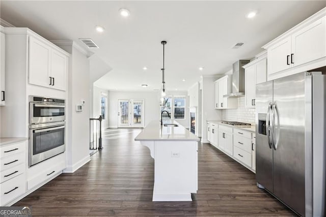 kitchen with sink, stainless steel appliances, wall chimney range hood, decorative light fixtures, and a kitchen island with sink