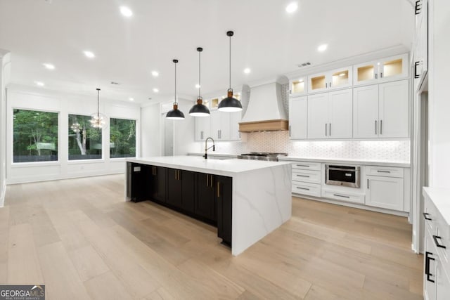 kitchen with white cabinetry, hanging light fixtures, light hardwood / wood-style floors, a center island with sink, and custom exhaust hood