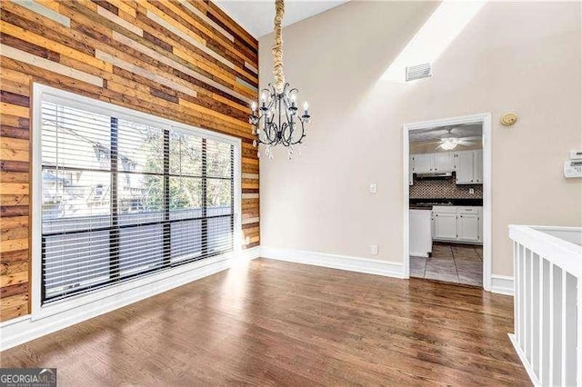 unfurnished dining area with ceiling fan with notable chandelier, wood walls, and dark hardwood / wood-style flooring