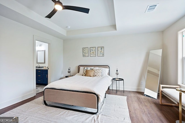 bedroom with a raised ceiling, visible vents, ensuite bath, wood finished floors, and baseboards