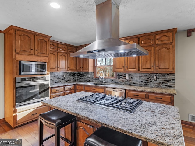 kitchen featuring appliances with stainless steel finishes, light stone counters, island range hood, sink, and a breakfast bar area