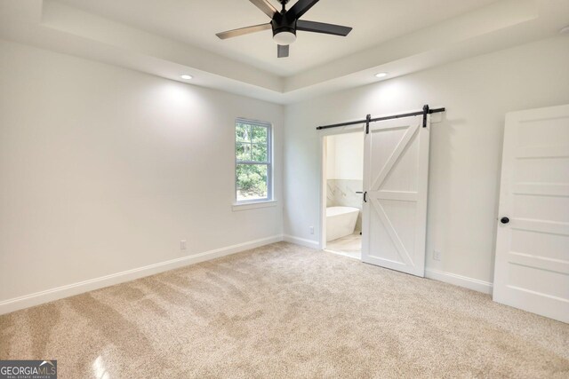 unfurnished bedroom with light carpet, ensuite bathroom, ceiling fan, a barn door, and a tray ceiling