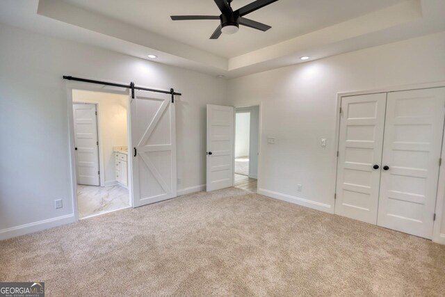 unfurnished bedroom with a raised ceiling, a barn door, ceiling fan, and light colored carpet