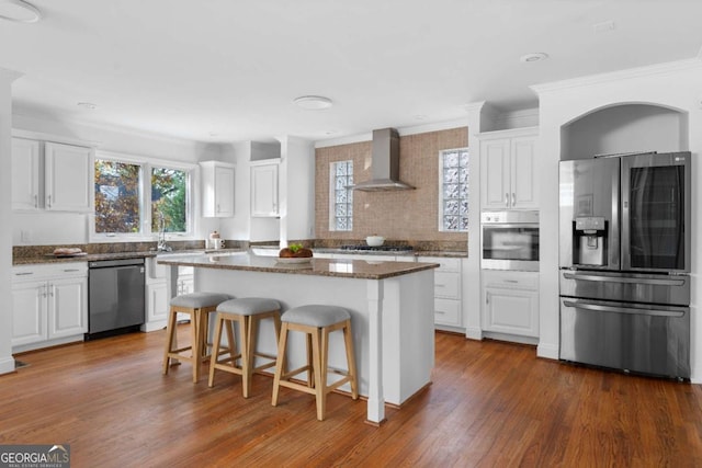 kitchen with dark stone counters, wall chimney range hood, appliances with stainless steel finishes, a kitchen island, and white cabinetry