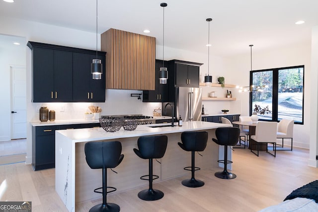 kitchen with sink, light hardwood / wood-style flooring, hanging light fixtures, high quality fridge, and an island with sink