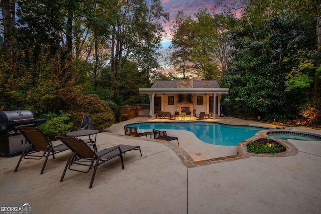 pool at dusk featuring an in ground hot tub, an outdoor structure, grilling area, a patio area, and an outdoor fireplace