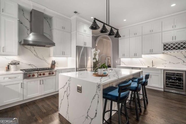 kitchen featuring a center island with sink, hanging light fixtures, wall chimney exhaust hood, stainless steel appliances, and beverage cooler