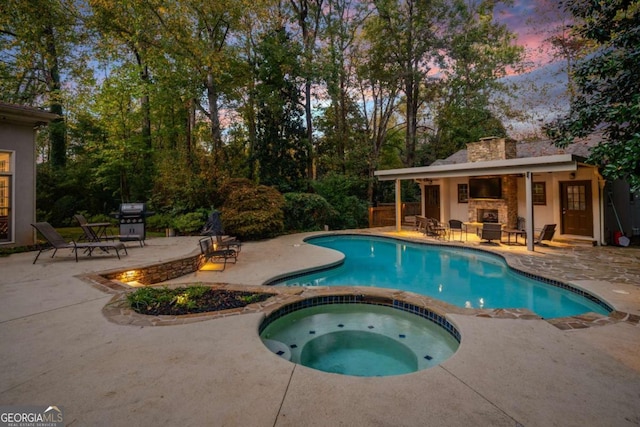 pool at dusk with a patio area, an in ground hot tub, grilling area, and exterior fireplace