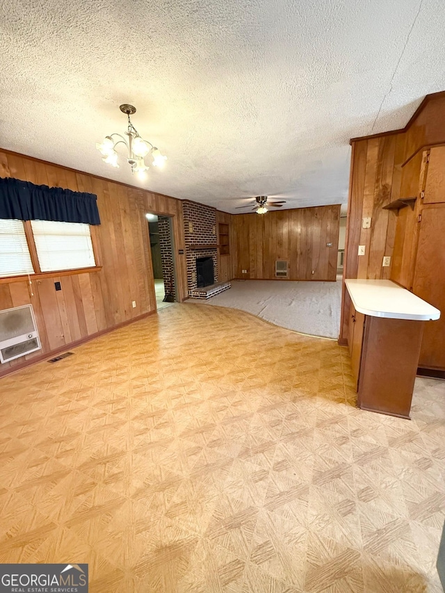 unfurnished living room featuring a textured ceiling, ceiling fan with notable chandelier, a brick fireplace, and wooden walls